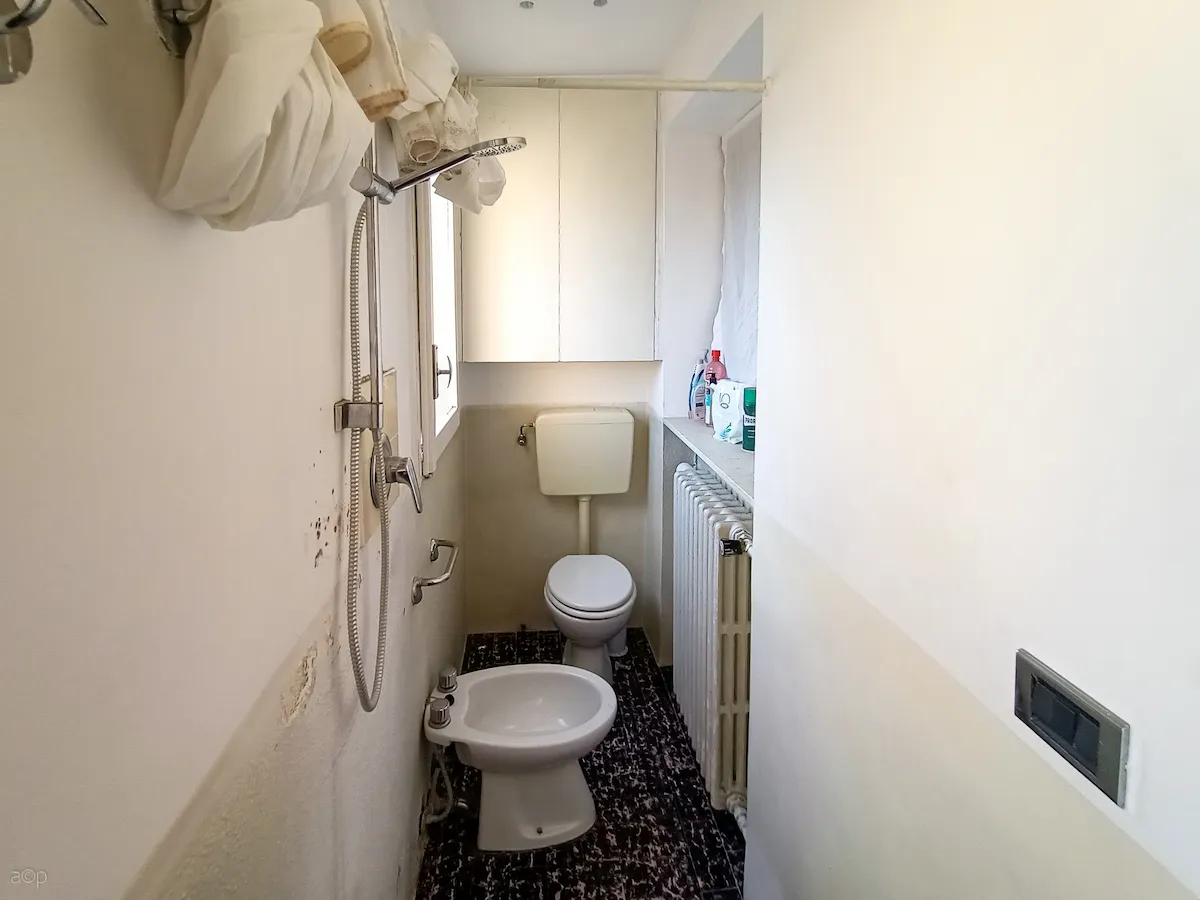 Fotografia di un bagno esistente molto stretto con pareti bianche rovinate, pavimento scuro maculato, un bidet e un WC con cassetta esterna, e un soffione doccia installato a parete senza box.
