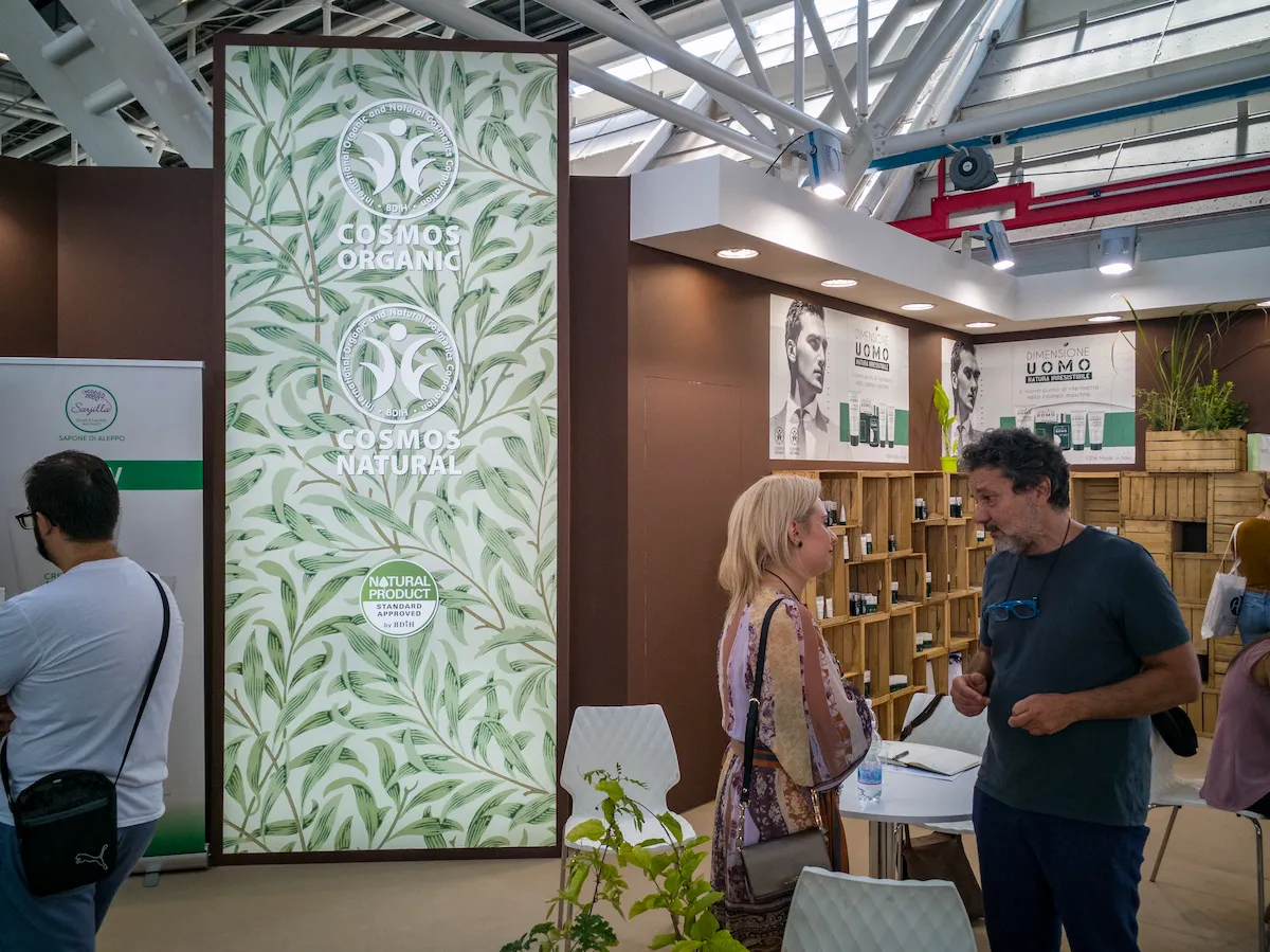 Stand fieristico ecosostenibile con scaffalature in legno grezzo tipo cassette della frutta e poster promozionali.