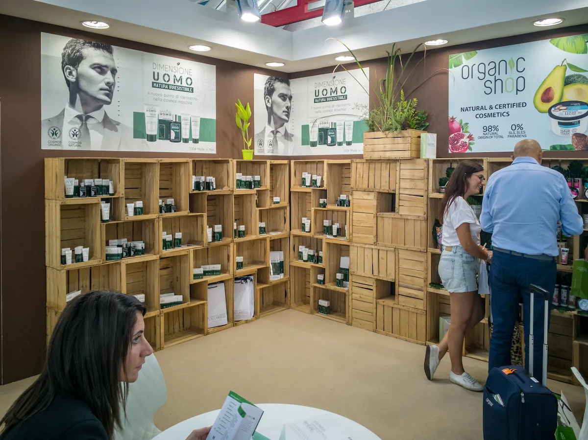 Stand fieristico ecosostenibile con scaffalature in legno grezzo tipo cassette della frutta e poster promozionali.