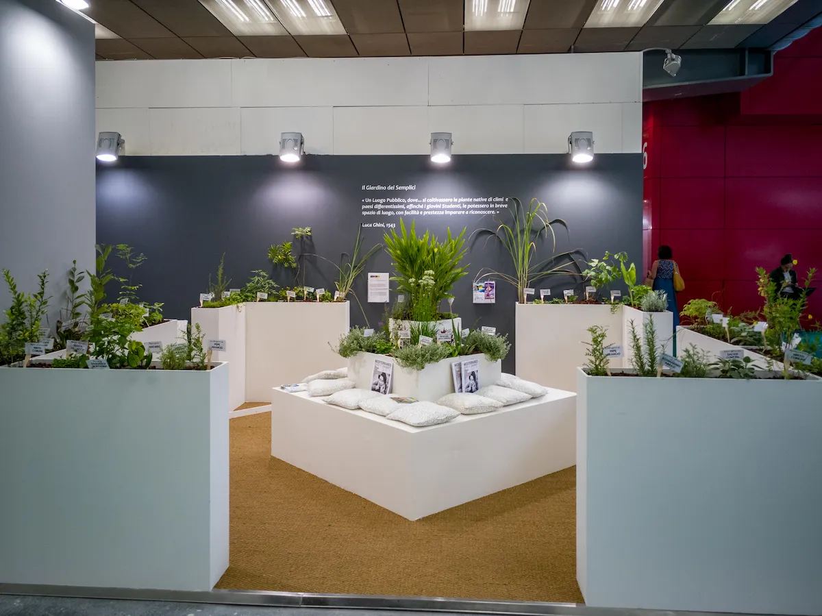 Allestimento interno con vasche bianche contenenti diverse varietà di piante vive e sedute a sacco.
